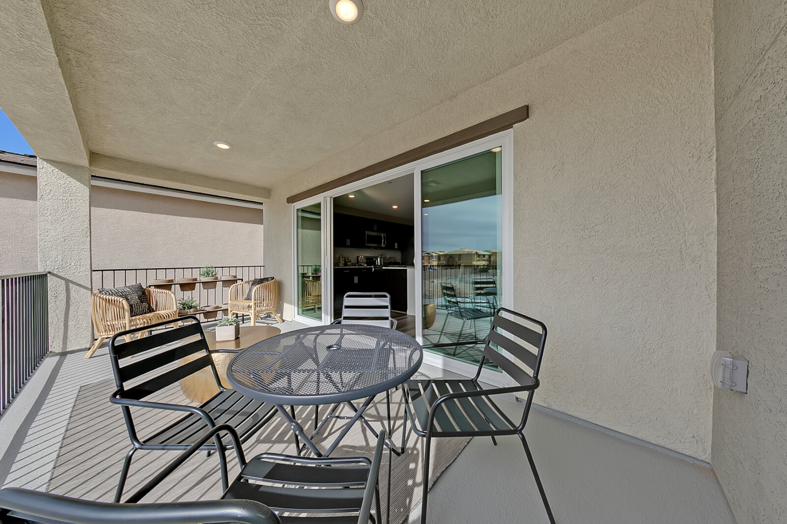 Covered balcony of an Indigo Plan 544 model home featuring a black metal patio dining set, woven accent chairs, and large sliding glass doors looking into a modern kitchen with dark cabinets.