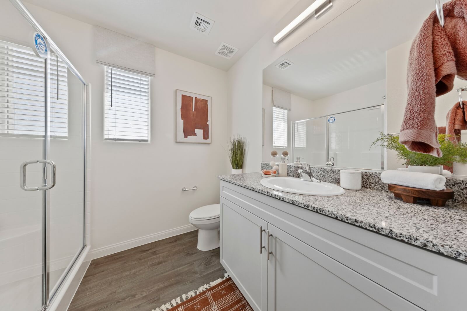 Luxurious primary bathroom with dark wood-look upgraded vinyl plank flooring, a grey granite double vanity, a walk-in glass shower, and a soaking tub under a window.