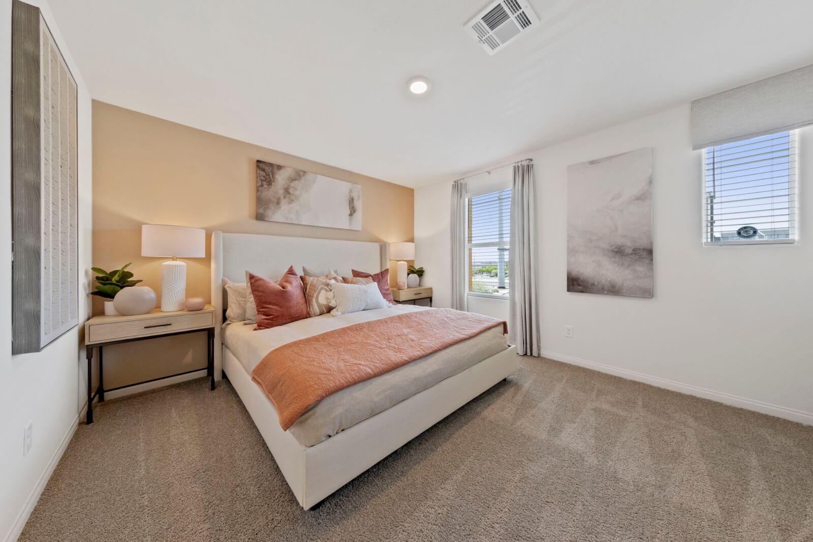 Primary bedroom with carpet and a soft peach accent wall featuring a wood-frame bed with neutral and rust bedding and a geometric mirror in a Solaris planned community home.