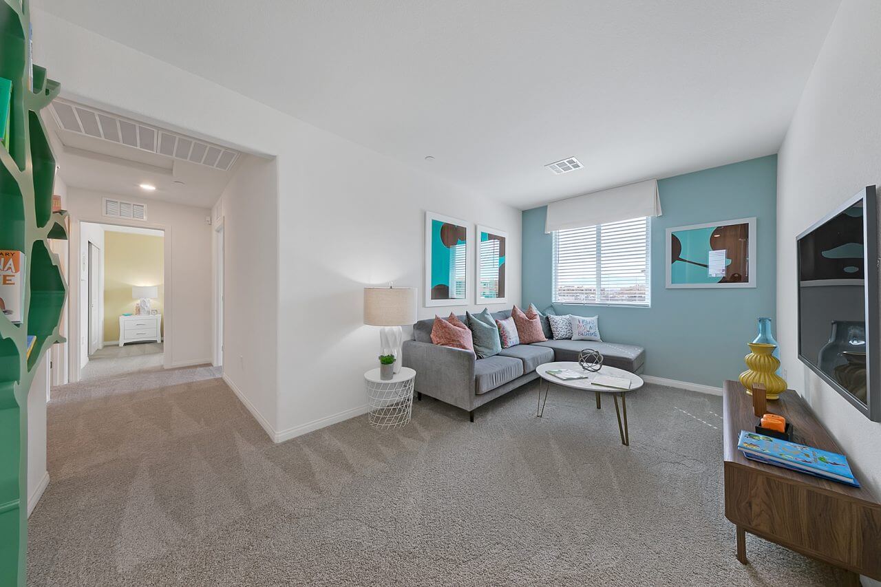 Carpeted loft lounge area in an Indigo Plan 541 model home at the Watercolor neighborhood, featuring a light blue accent wall, a grey sectional sofa, an oval white coffee table, and a glimpse of a green built-in bookshelf