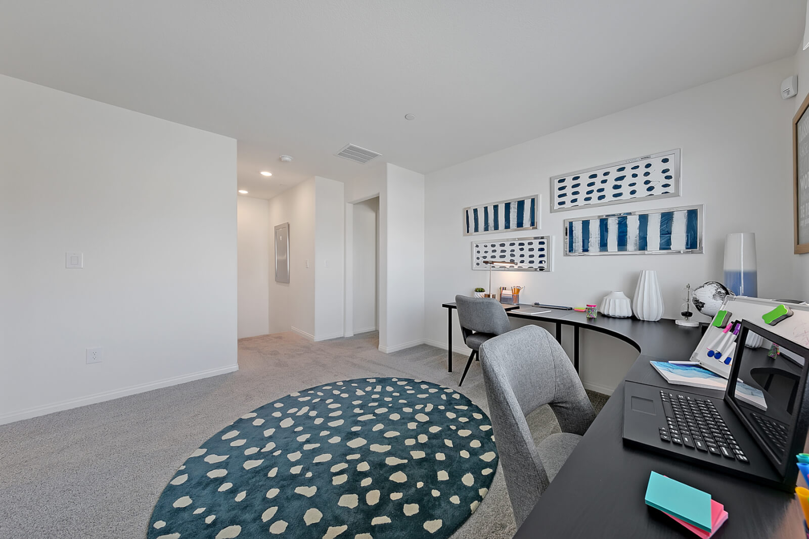 Carpeted loft space in a Crimson Plan 520 model home configured as a shared dual home office, featuring a curved black wrap-around desk, two grey upholstered office chairs, a laptop, and a large round blue patterned area rug.